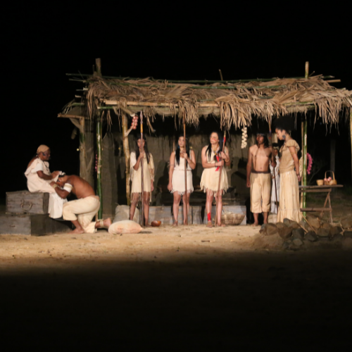 Cena de uma peça de teatro encenada durante a noite em uma área aberta. Os atores estão caracterizados como índios e escravos.