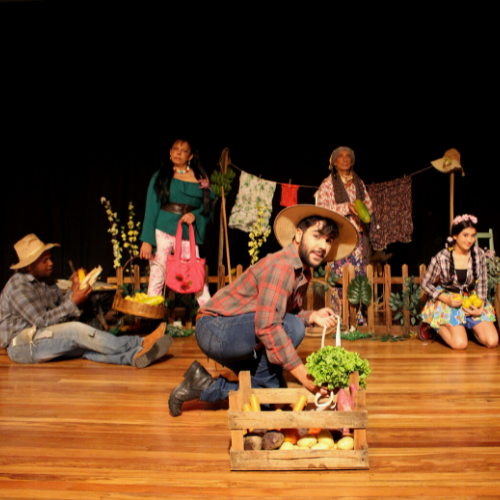 Grupo de atores em um palco caracterizados em seus personagens. Alguns usam chapéu. Em destaque, há um ator que segura uma verdura.